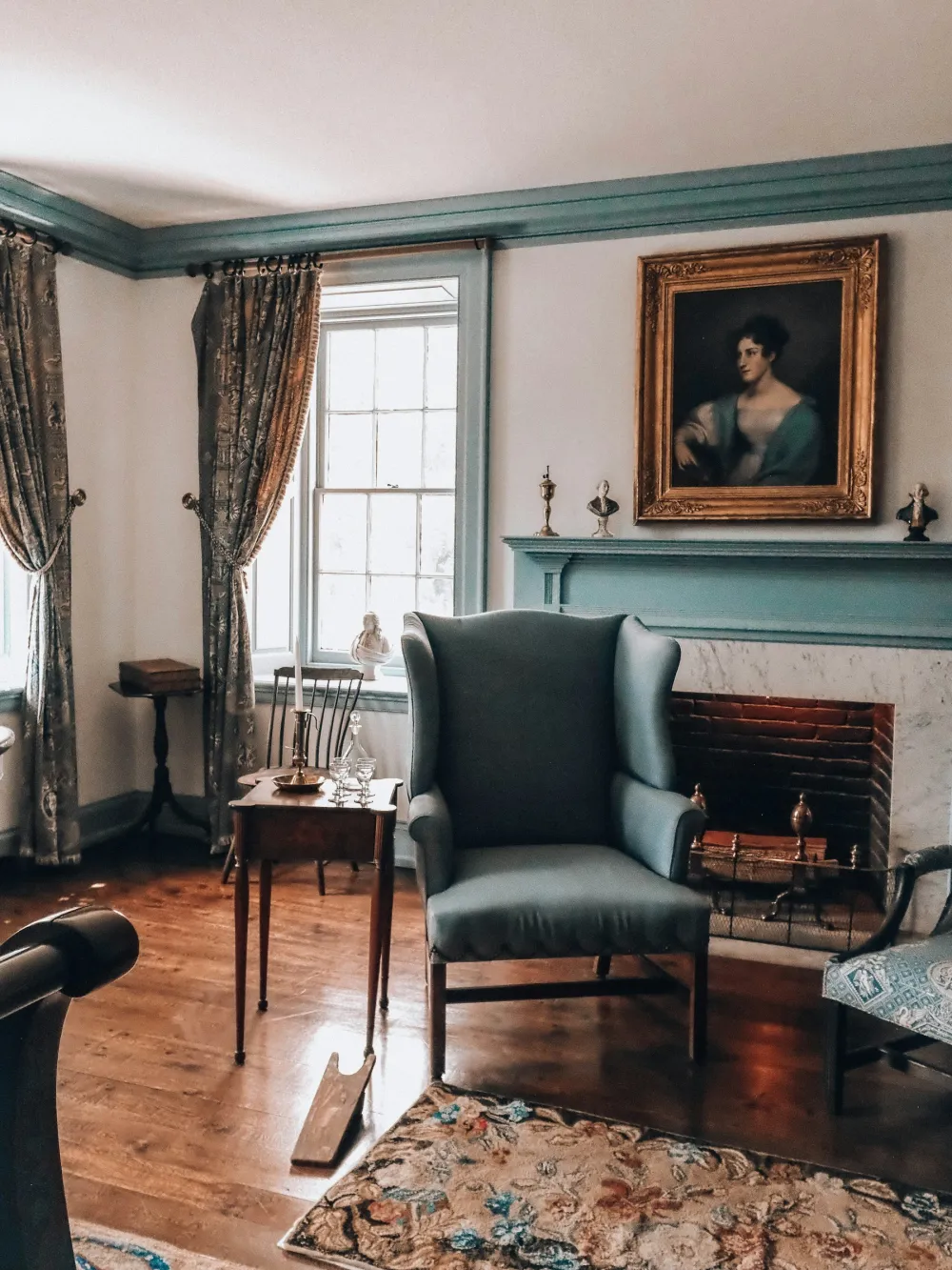 A period sitting room with a painted fireplace surround, a wing chair and a sash window